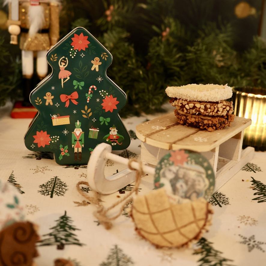 Boîte verte en forme de sapin décoré, accompagnée de biscuits présentés sur une luge.
