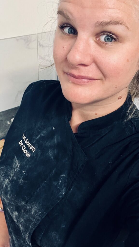 Selfie en plan poitrine d'Ophélie, alias Choue, une femme blonde aux yeux bleus, souriante et regardant directement l'objectif. Elle porte une veste de biscuitière noire, visiblement couverte de farine blanche, avec l'inscription brodée en blanc 'Les Secrets De Choue' sur le côté gauche. L'image est prise en selfie, en plan rapproché, mettant en avant son visage et le haut de son uniforme de travail, suggérant une activité de pâtisserie authentique et passionnée.